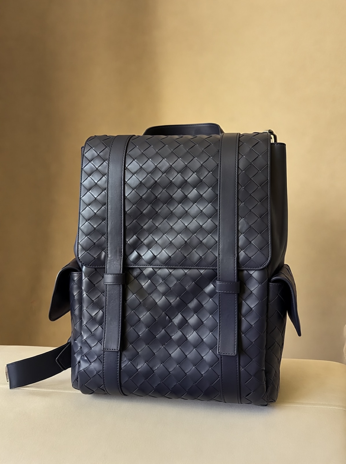 A navy woven leather backpack from the front on a cream seat against a warm wall