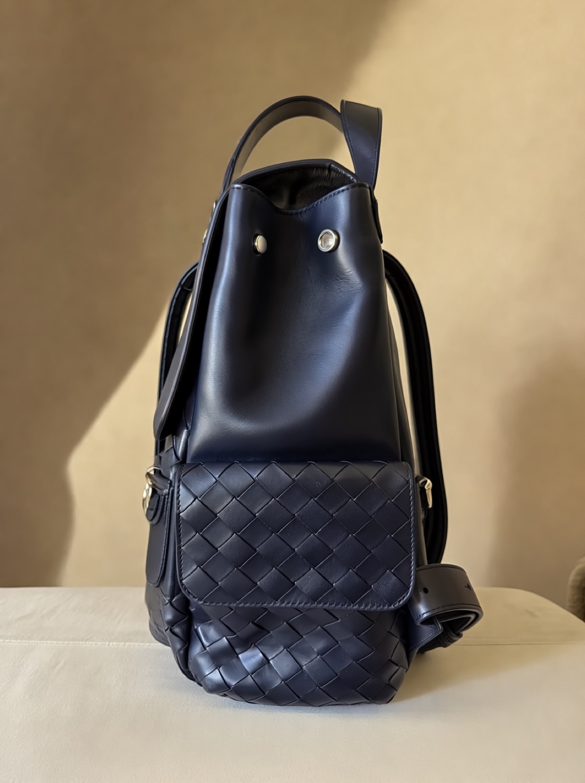 The backpack from the side, showing the smooth leather side panel and woven side pocket