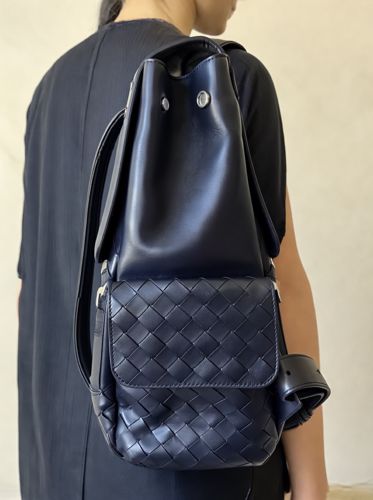 A navy woven leather backpack worn over the shoulder by a person in a black dress, face out of frame