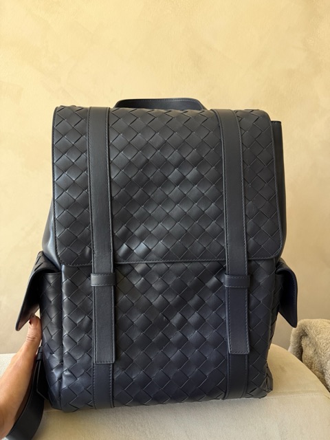 The starting photo: a navy woven leather backpack standing on a cream chair, held by the owner's hand, taken on a phone