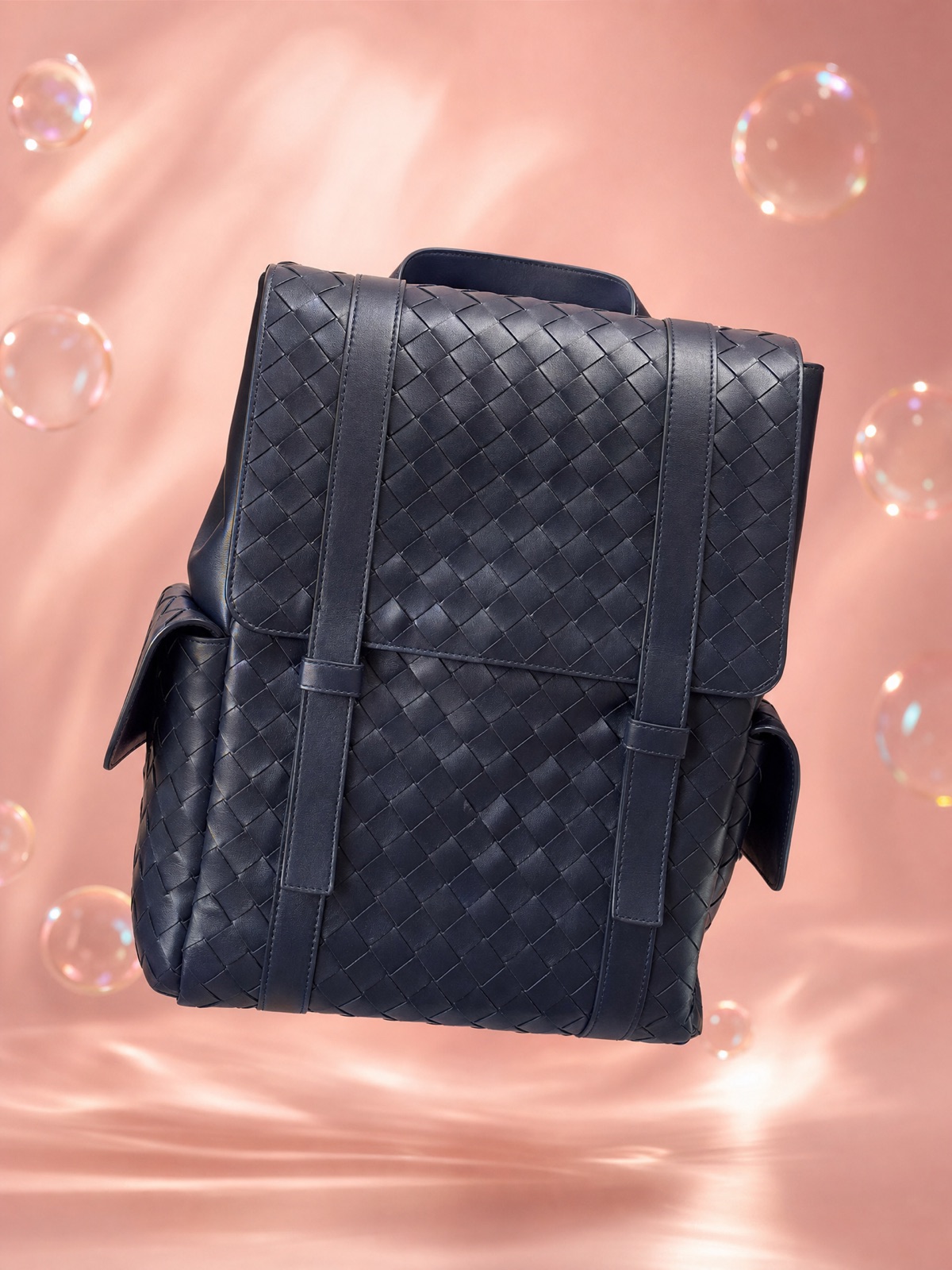 A navy woven leather backpack floating on warm rose-pink with soft bubbles