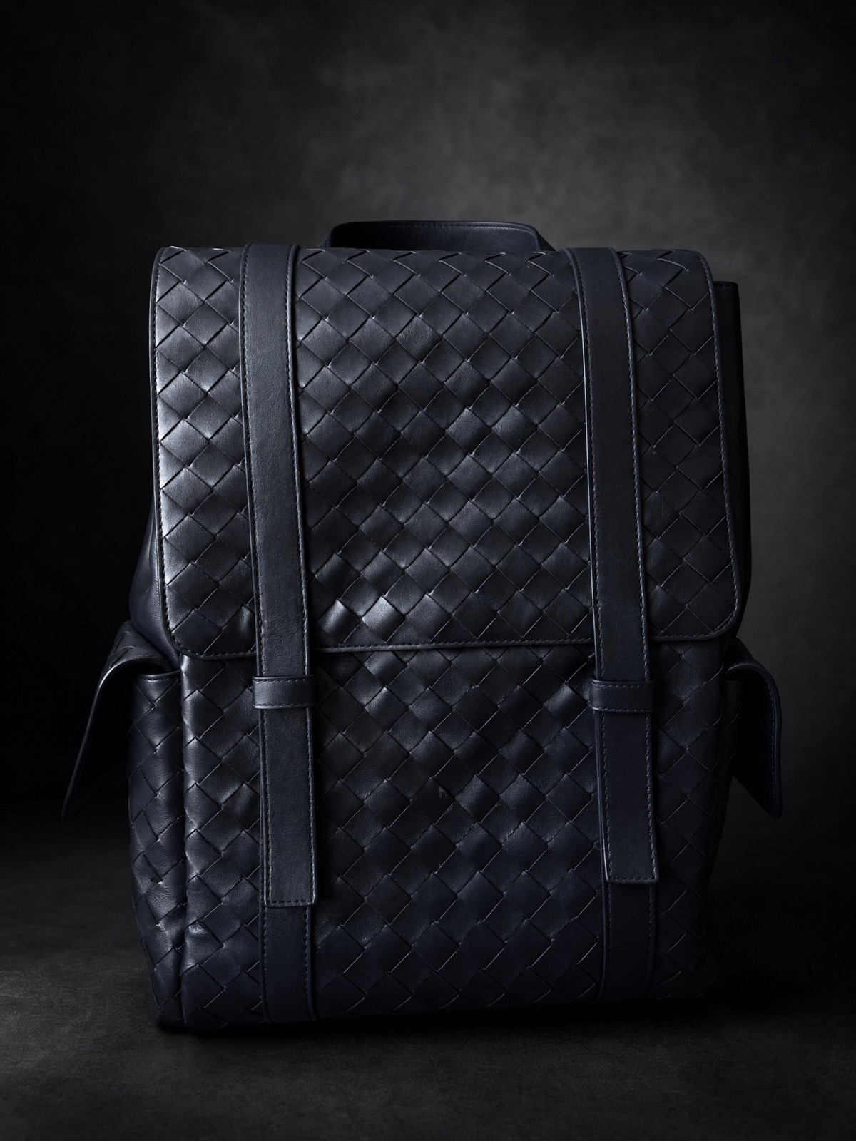 A navy woven leather backpack against a dark backdrop under a single dramatic light