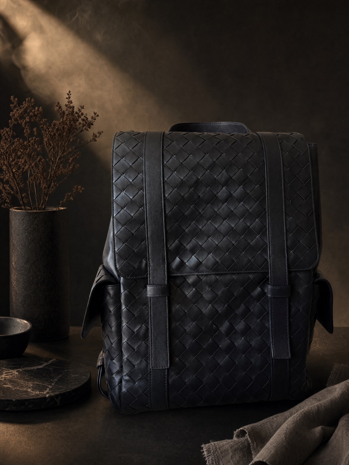 The backpack on a dark table with dried grasses and a linen cloth, warm light from the left