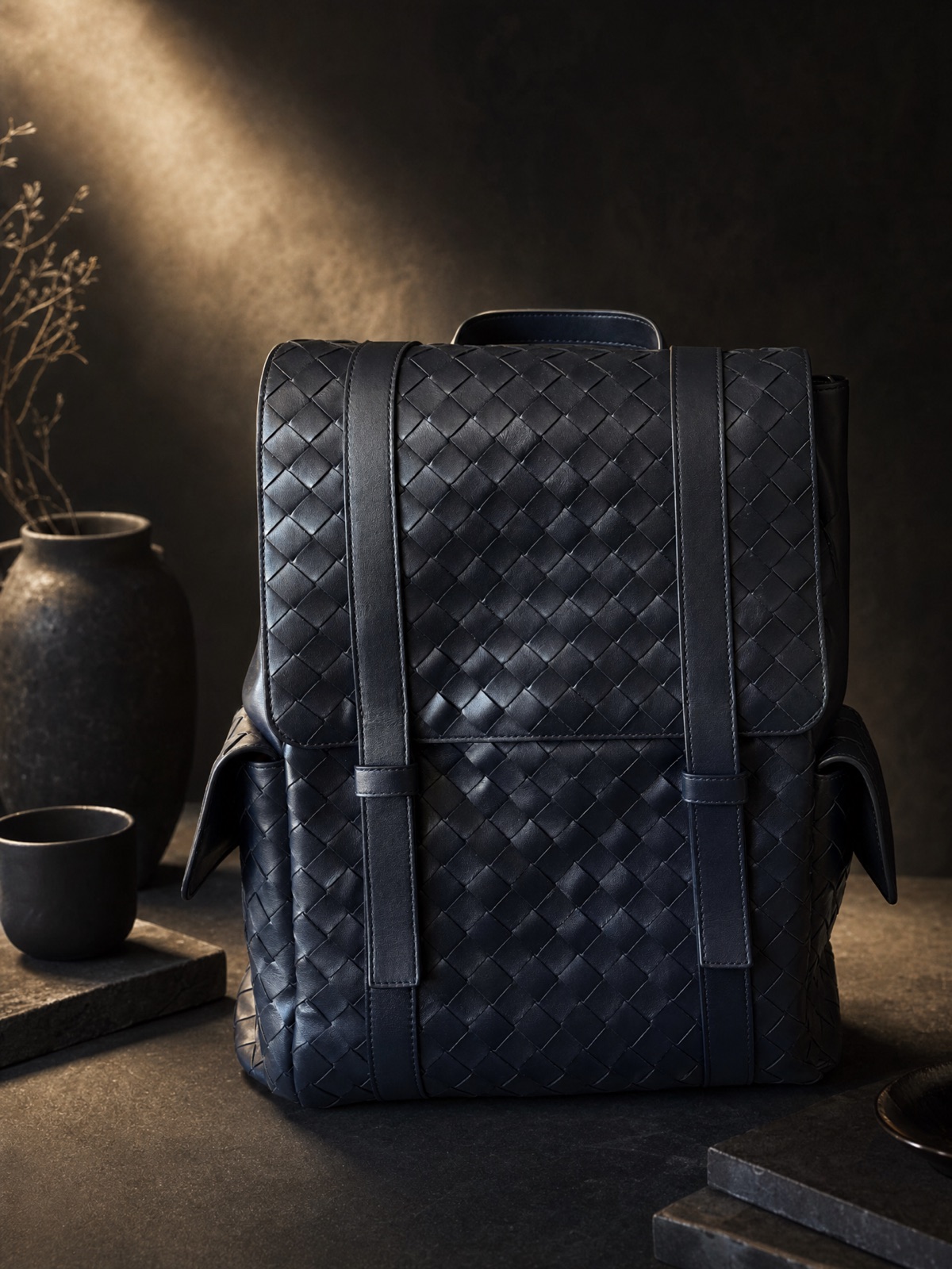 A navy woven leather backpack on a dark table beside a stone vase and cup, warm side light