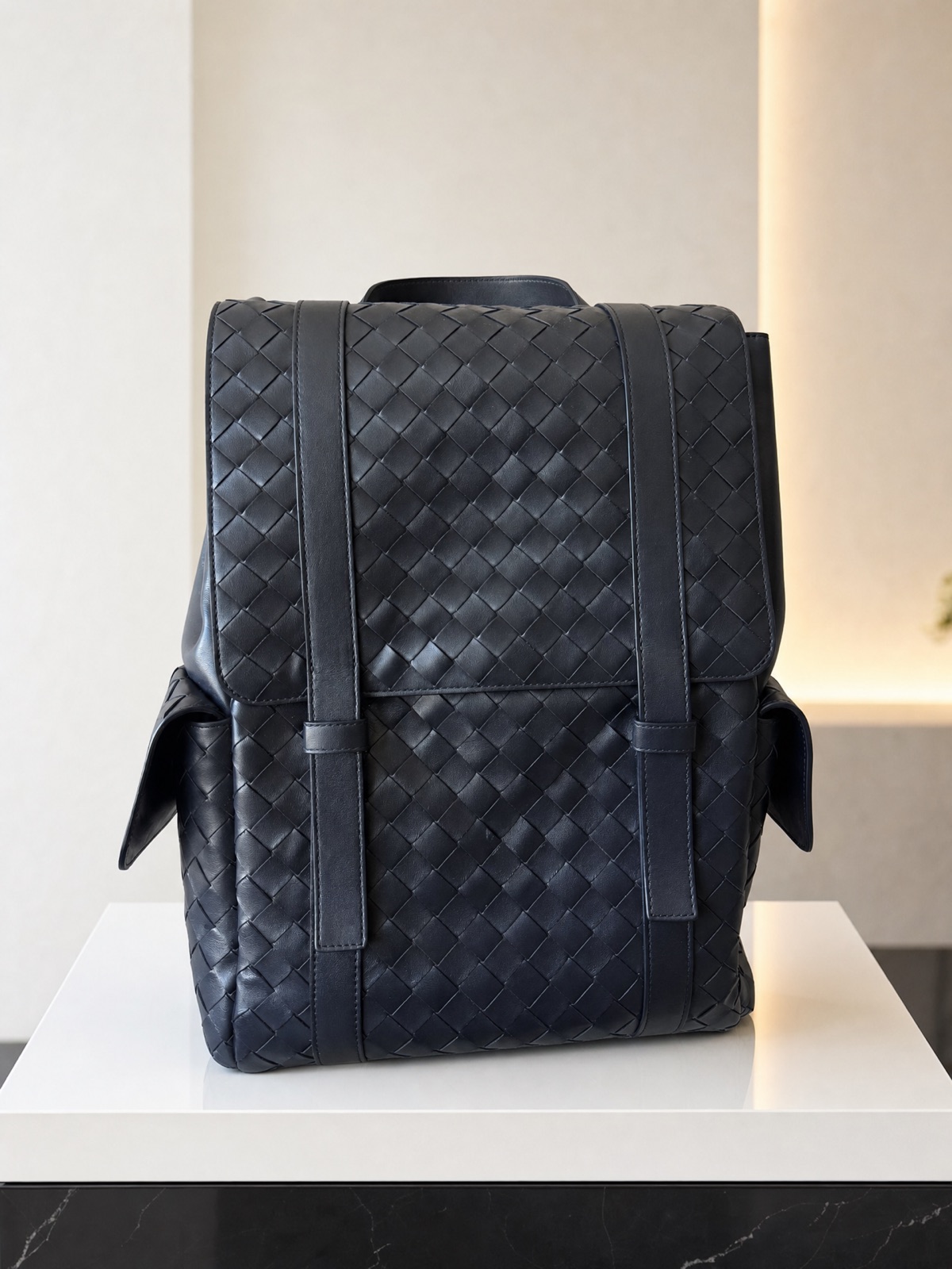 A navy woven leather backpack on a white plinth in a calm, minimal room