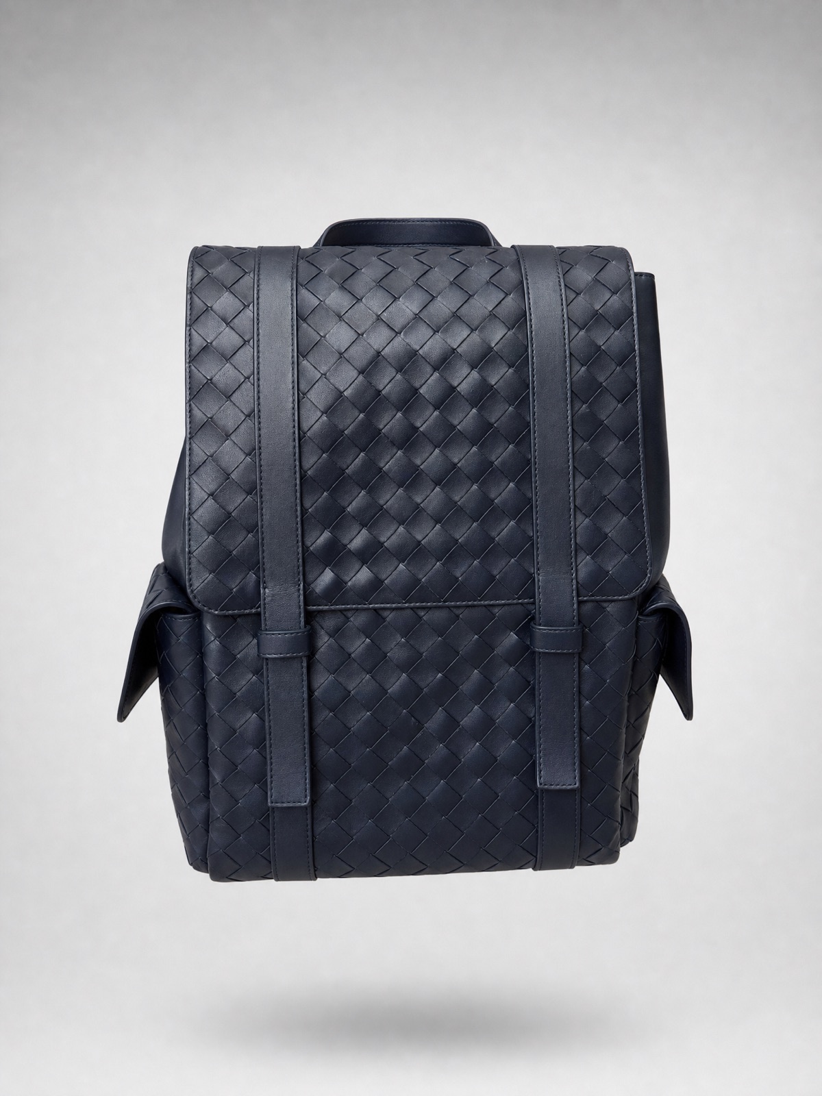 A navy woven leather backpack floating dead-centre on soft grey, crisp and clean